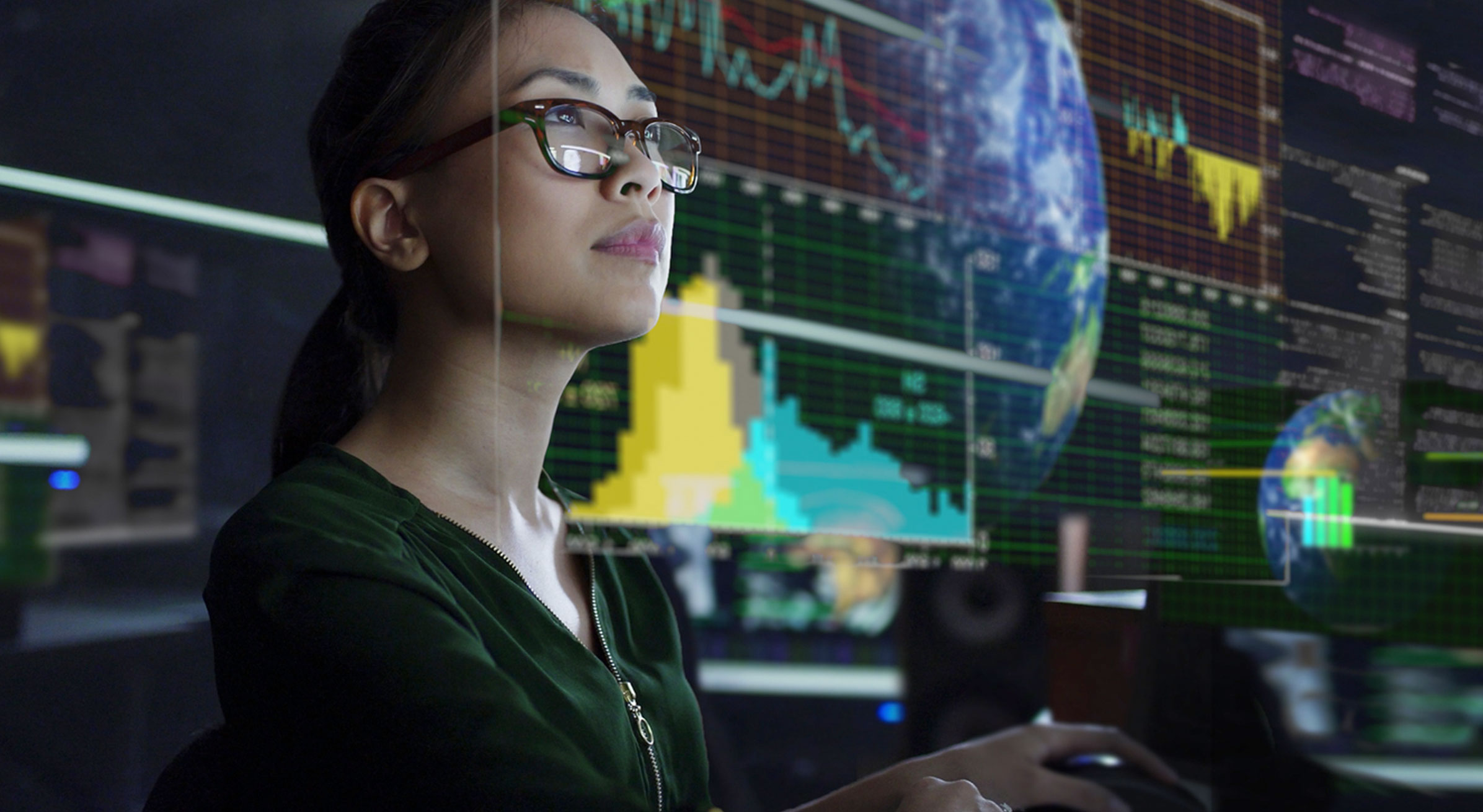 A person wearing glasses focused on multiple digital screens displaying data visualizations and charts, surrounded by a high-tech environment with globes and detailed graphs in the background.
